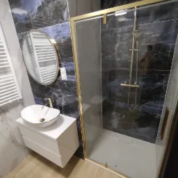 Elegancka łazienka po remoncie: kabina prysznicowa ze złotymi ramami, umywalka nablatowa na białej szafce, okrągłe lustro i marmurowe płytki w odcieniach granatu.