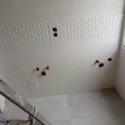 Ściana łazienki z ułożonymi białymi płytkami, dekoracyjnym pasem z motywem liści i wyprowadzonymi instalacjami wodno-kanalizacyjnymi, widok z góry ze schodów drabiny.