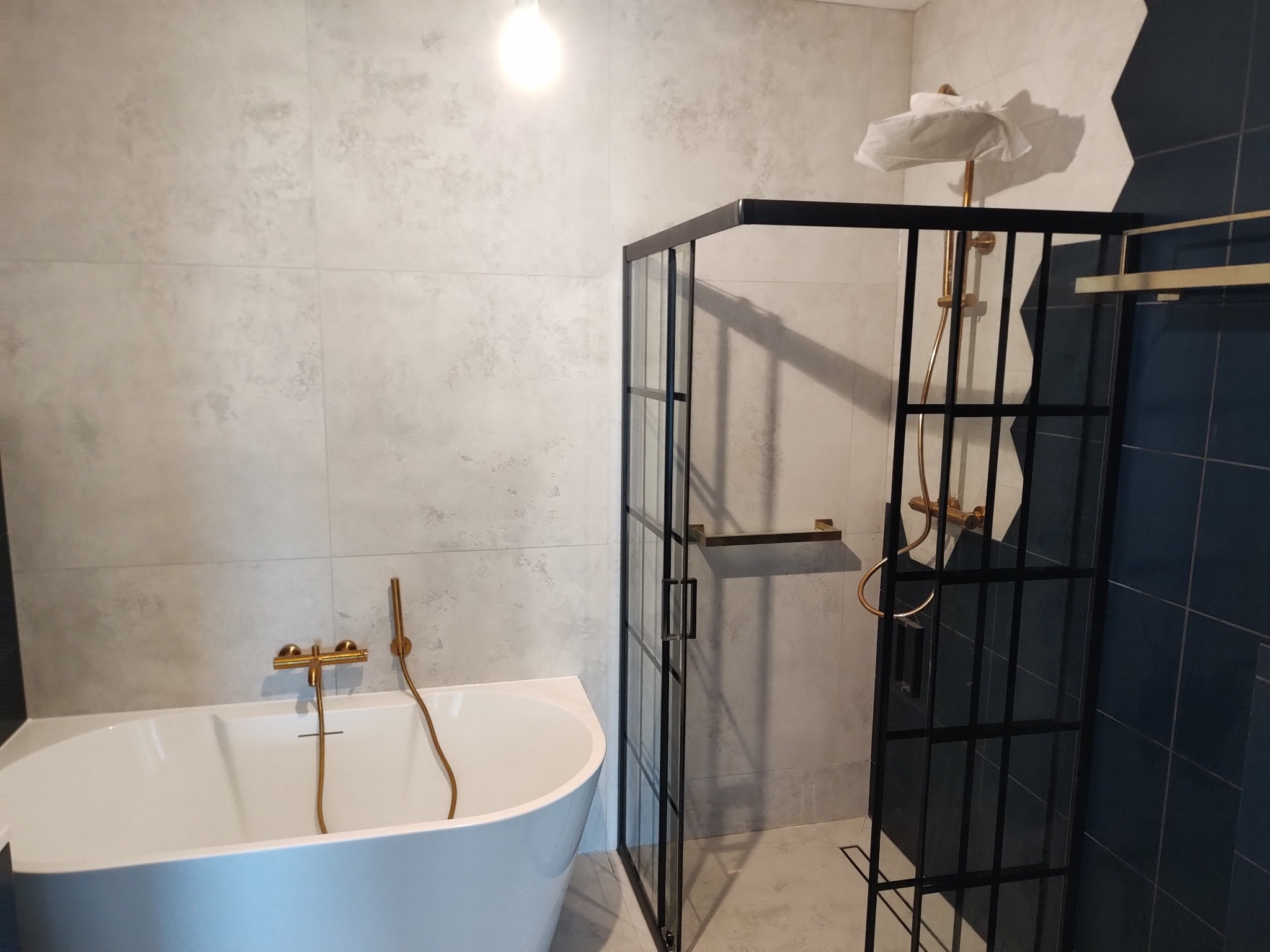 Nowoczesna łazienka z białą wanną, złotą armaturą i kabiną prysznicową z czarnymi profilami. Jasne płytki na ścianach i podłodze.