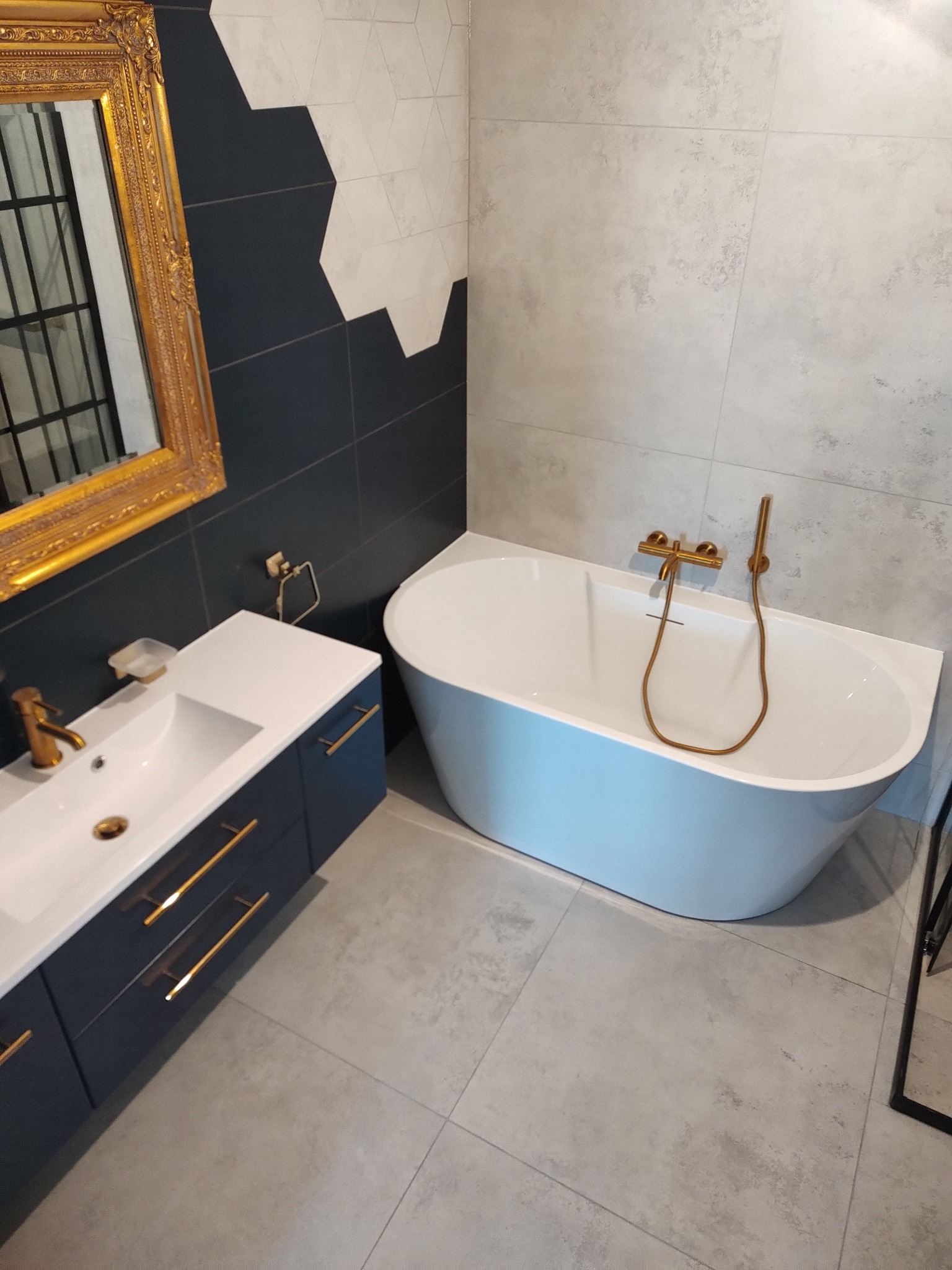 Elegancka łazienka: wolnostojąca wanna, umywalka z szafką w kolorze granatowym, złote detale armatury i dekoracyjne lustro w złotej ramie. Nowoczesne płytki na ścianach i podłodze.
