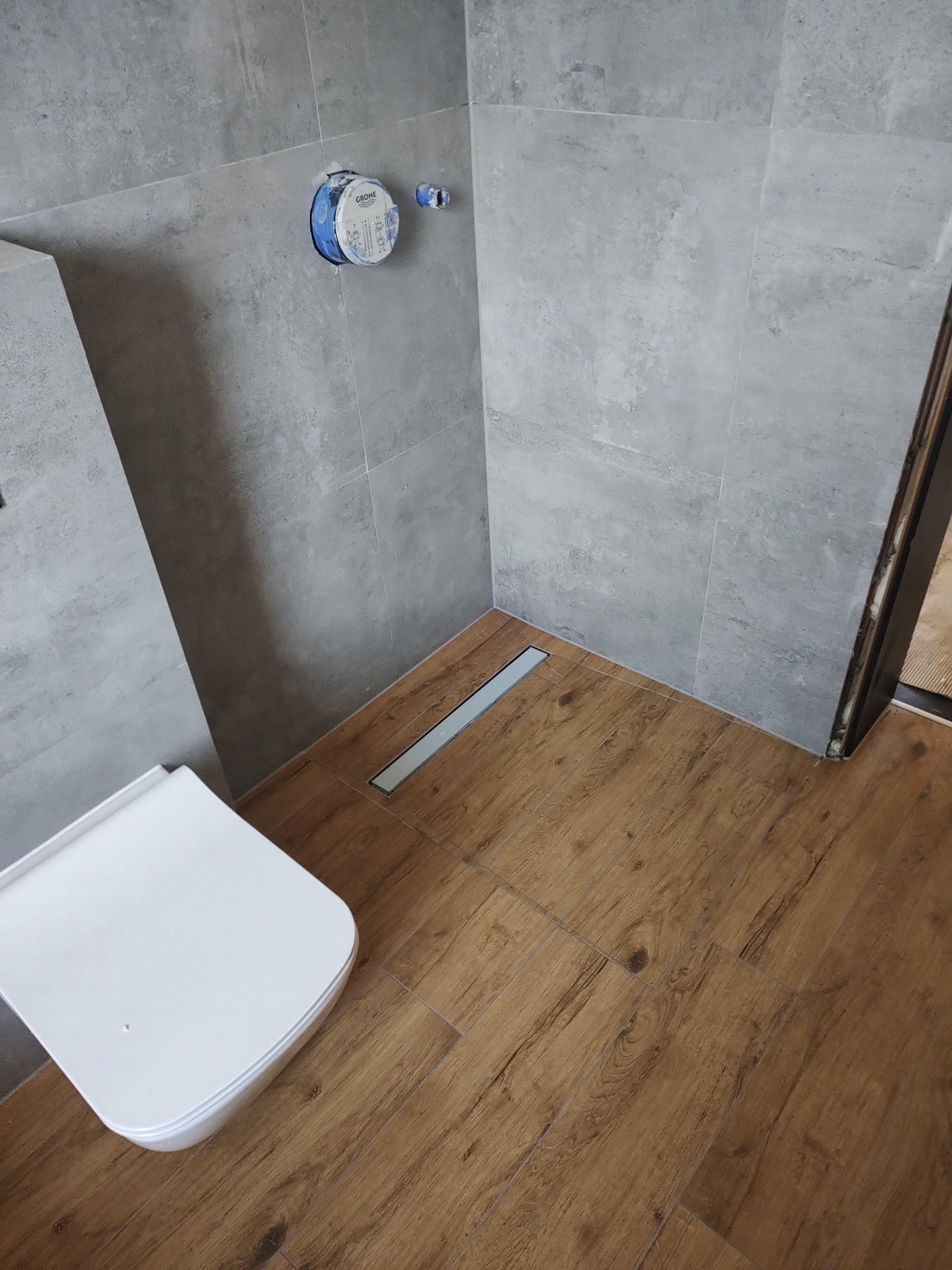 Nowoczesna łazienka z szarymi płytkami ściennymi i drewnopodobną podłogą. Widoczny biały sedes i odpływ liniowy. Minimalistyczny design i staranne wykończenie.