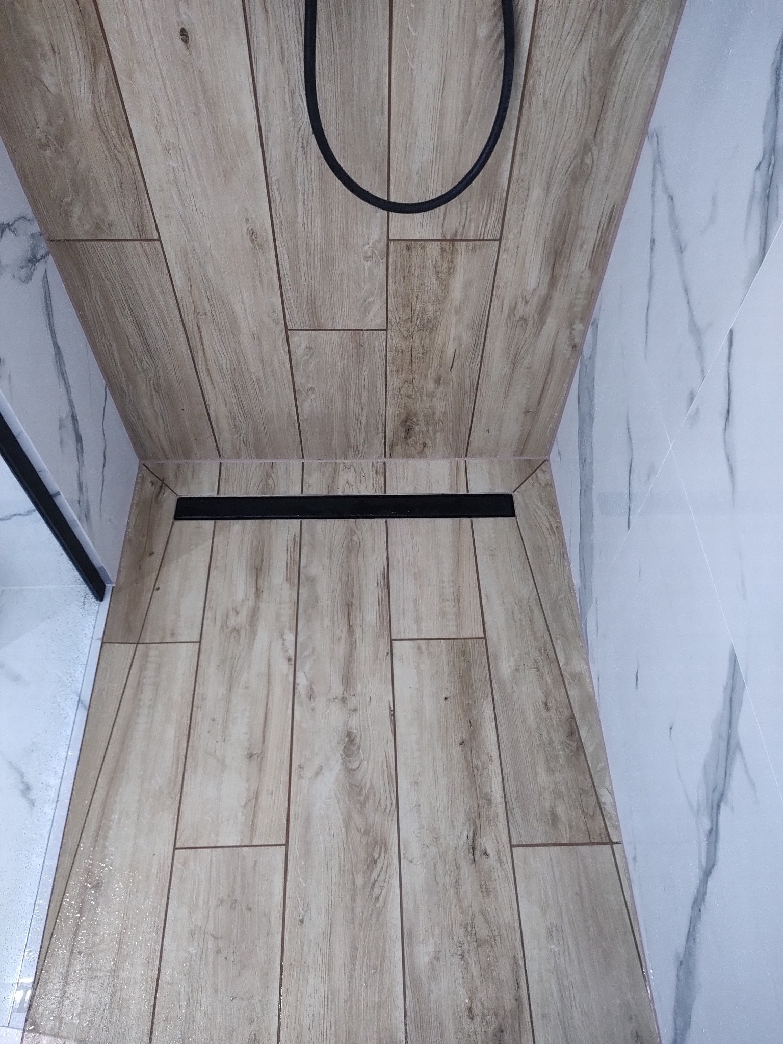 Wykończona kabina prysznicowa z odpływem liniowym. Płytki imitujące drewno na podłodze, marmurowe płytki na ścianach. Czarny wąż prysznicowy w górnej części kadru.