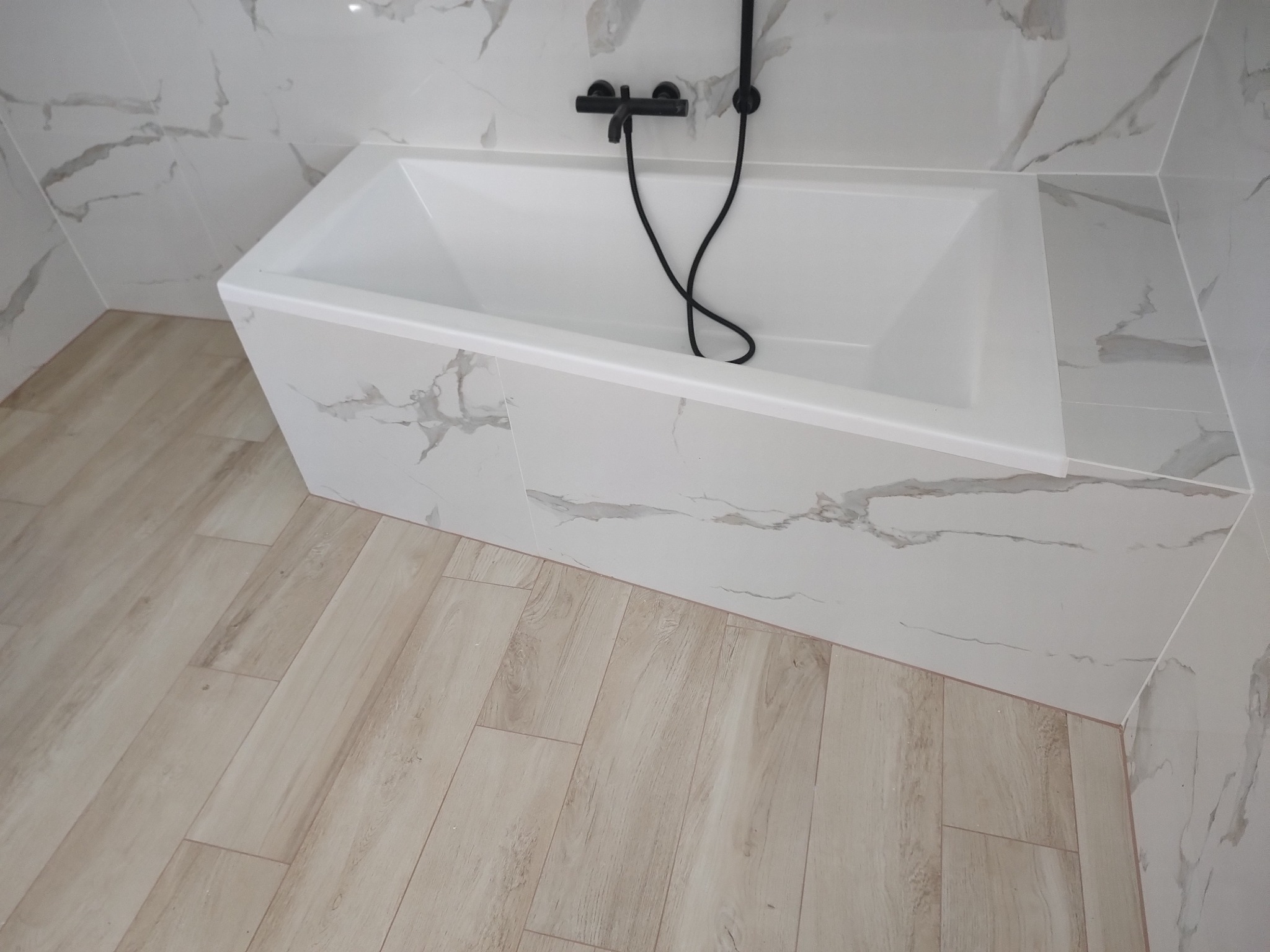 Biała wanna z czarną armaturą w łazience wykończonej płytkami imitującymi marmur i drewno. Minimalistyczny design i nowoczesne wykończenie.