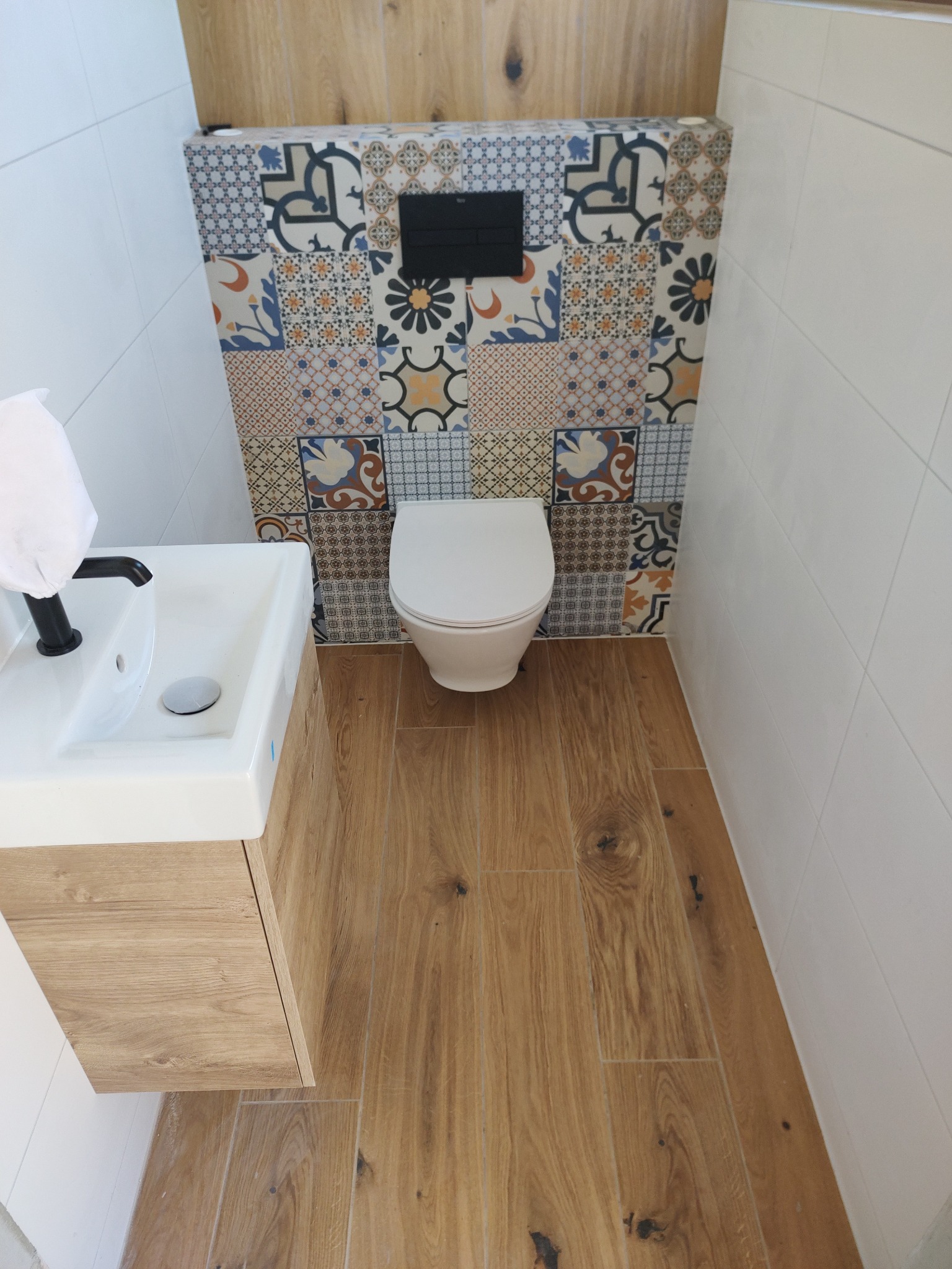 Nowoczesna łazienka po remoncie: toaleta, umywalka z szafką, drewnopodobna podłoga i ściana z mozaikowych płytek nad toaletą.