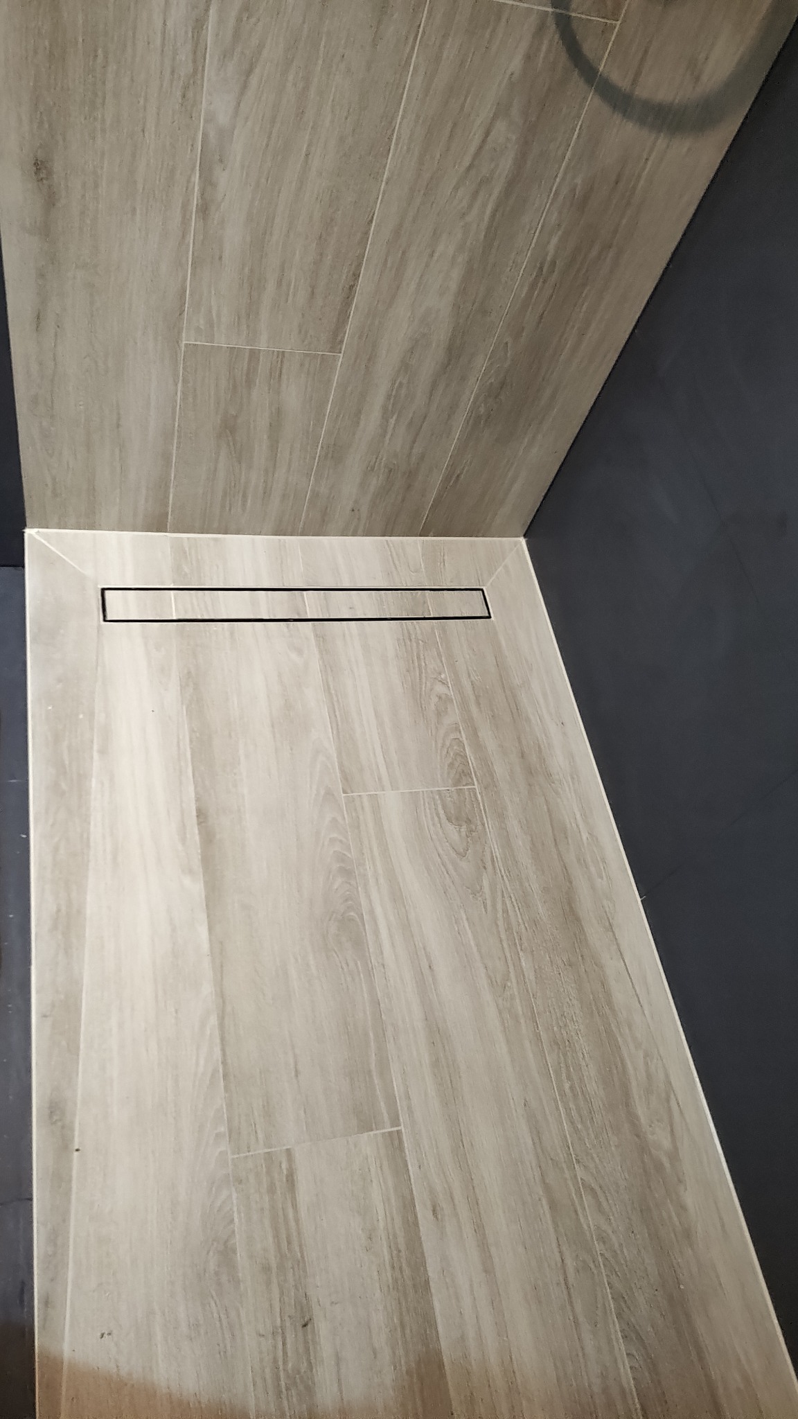 Beżowe płytki imitujące drewno w kabinie prysznicowej z odpływem liniowym. Ciemnoszara ściana. Ujęcie z góry.