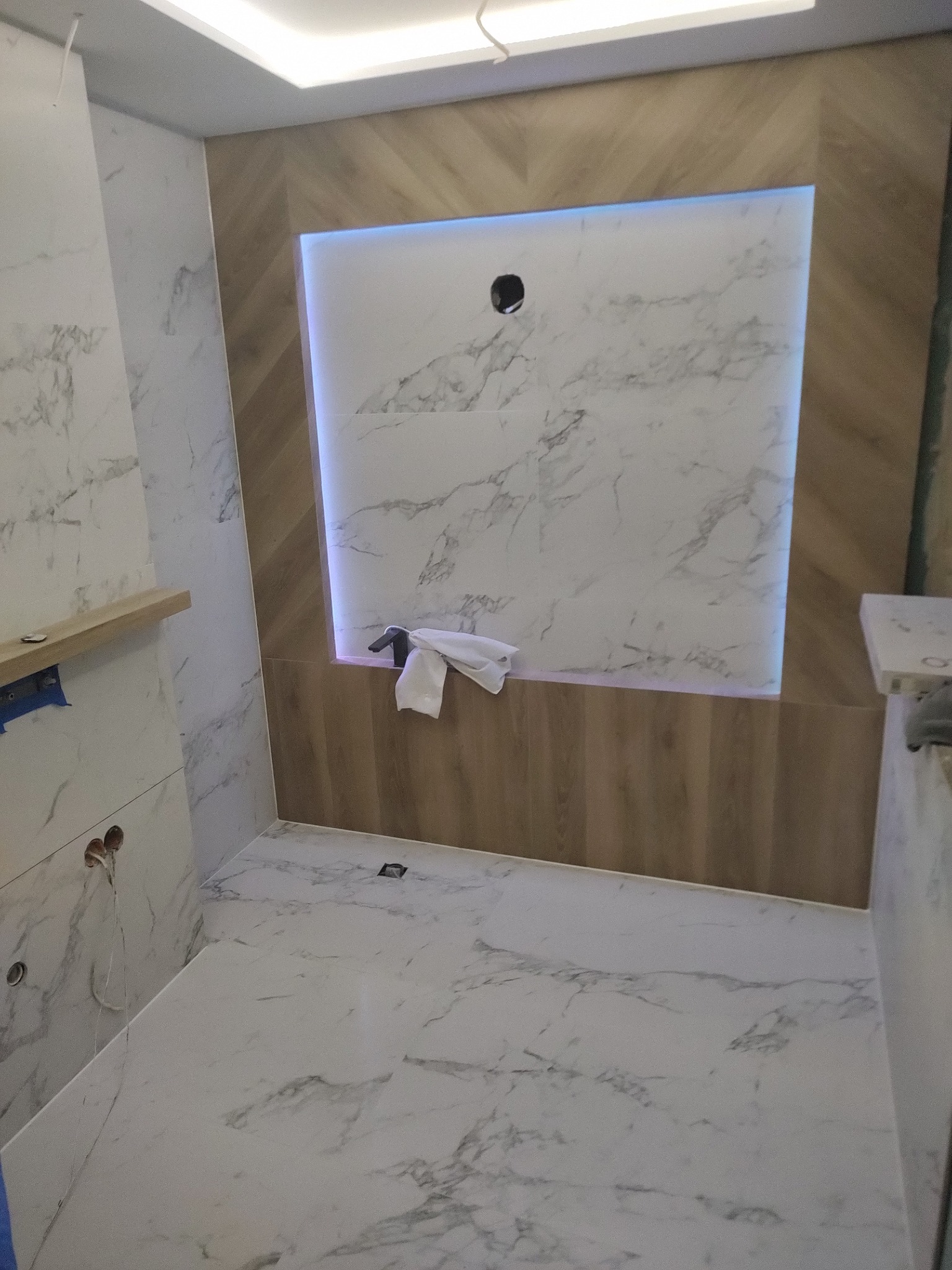 Nowoczesna łazienka z białymi płytkami imitującymi marmur i drewnianymi akcentami, podświetlona listwą LED, w trakcie wykańczania.