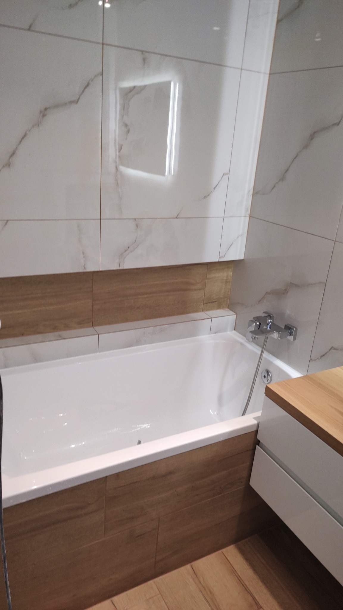 Biała wanna z obudową z płytek imitujących drewno, szare płytki ścienne z subtelnym wzorem marmuru i podświetlane lustro w wykończonej łazience.