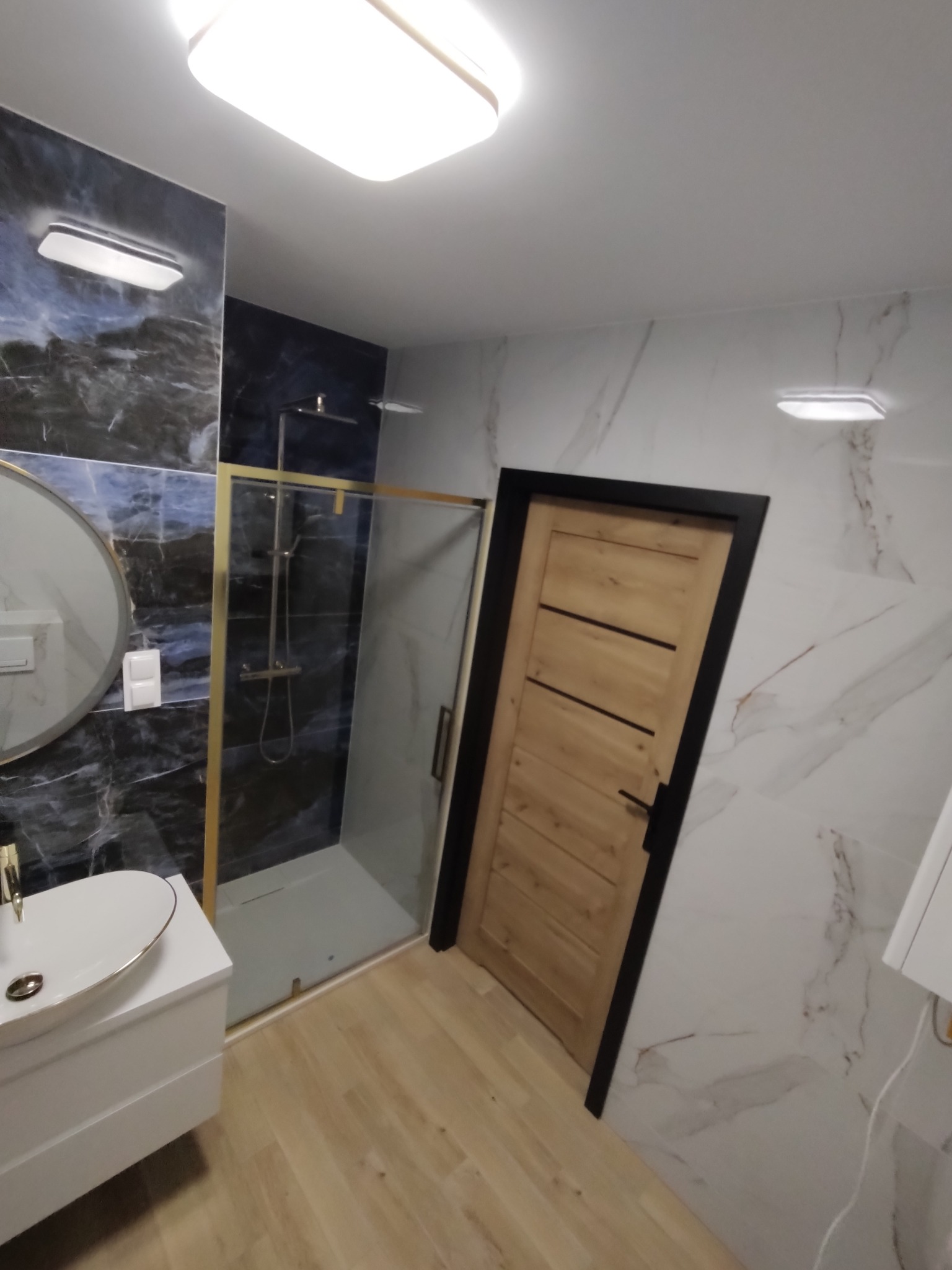 Nowoczesna łazienka po remoncie: kabina prysznicowa ze złotymi elementami, umywalka z szafką i drewniane drzwi z czarną ościeżnicą na tle marmurowych płytek.