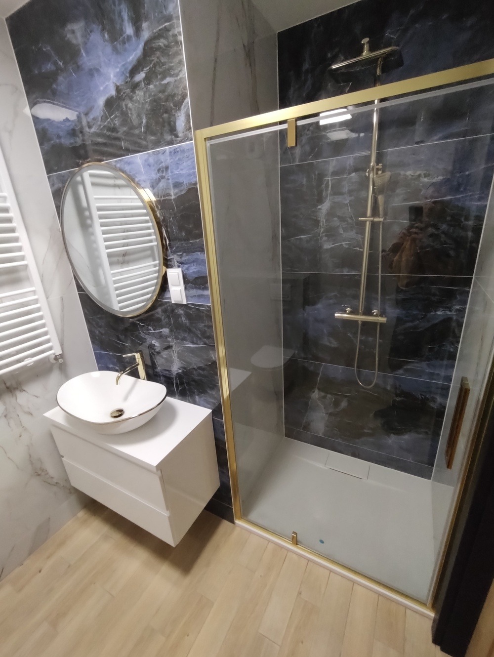 Elegancka łazienka po remoncie: kabina prysznicowa ze złotymi ramami, umywalka nablatowa na białej szafce, okrągłe lustro i marmurowe płytki w odcieniach granatu.