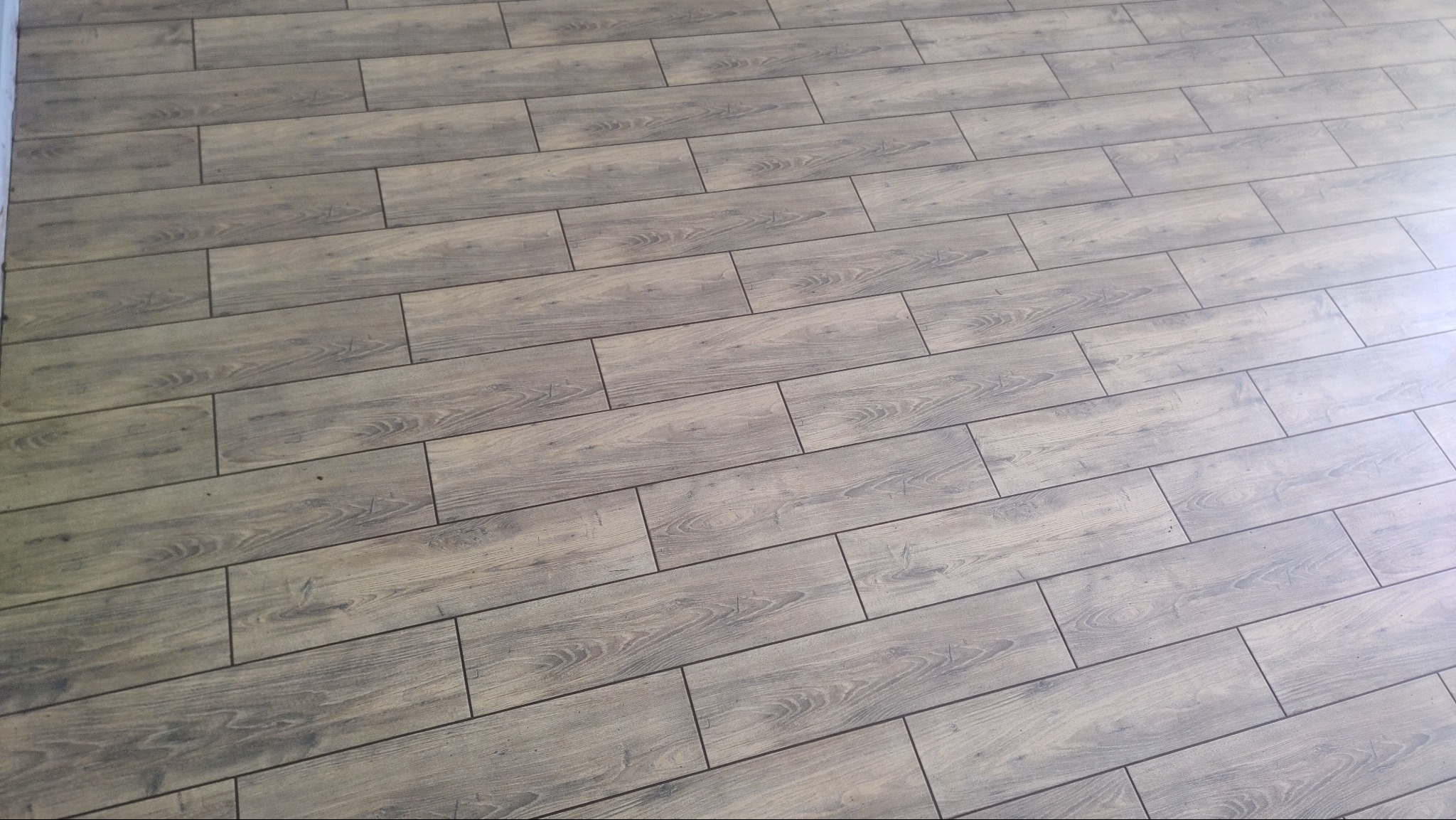 Ukończona podłoga z paneli imitujących drewno, ułożona pod kątem. Widoczne delikatne wzory słojów i fugi. Oświetlenie z prawej strony dodaje jasności.