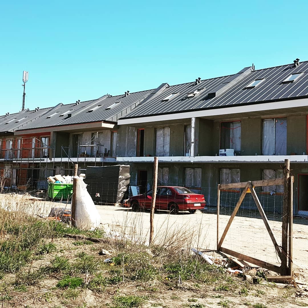 Rząd nowych domów w trakcie budowy z szarą elewacją i ciemnymi dachami, widoczne rusztowania, śmieci i czerwony samochód na pierwszym planie.