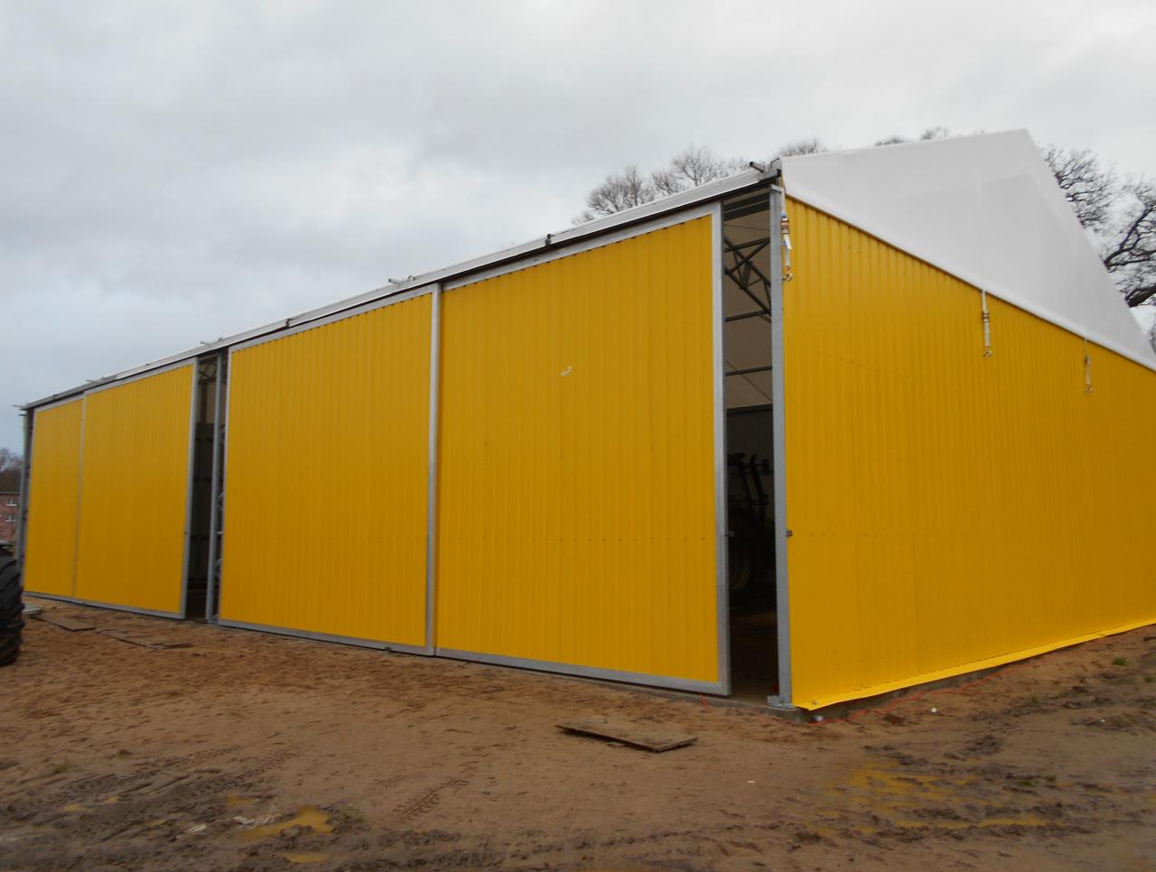 Żółty magazyn z płyt warstwowych z przesuwnymi drzwiami, widok z zewnątrz na błotnistym terenie.