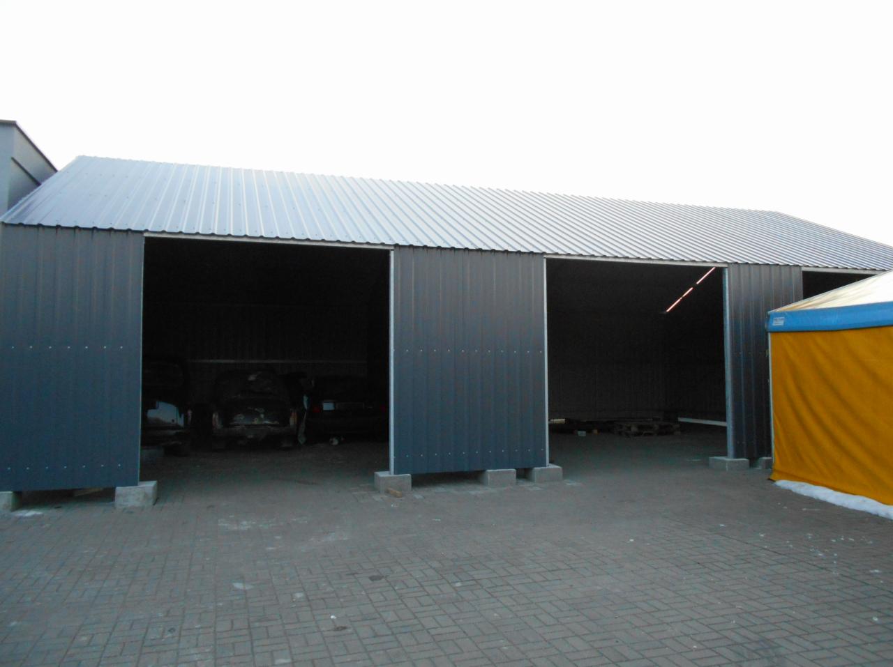 Szeroki, szary garaż blaszany z trzema otwartymi wrotami, widoczne betonowe fundamenty i częściowo zaparkowane samochody wewnątrz.