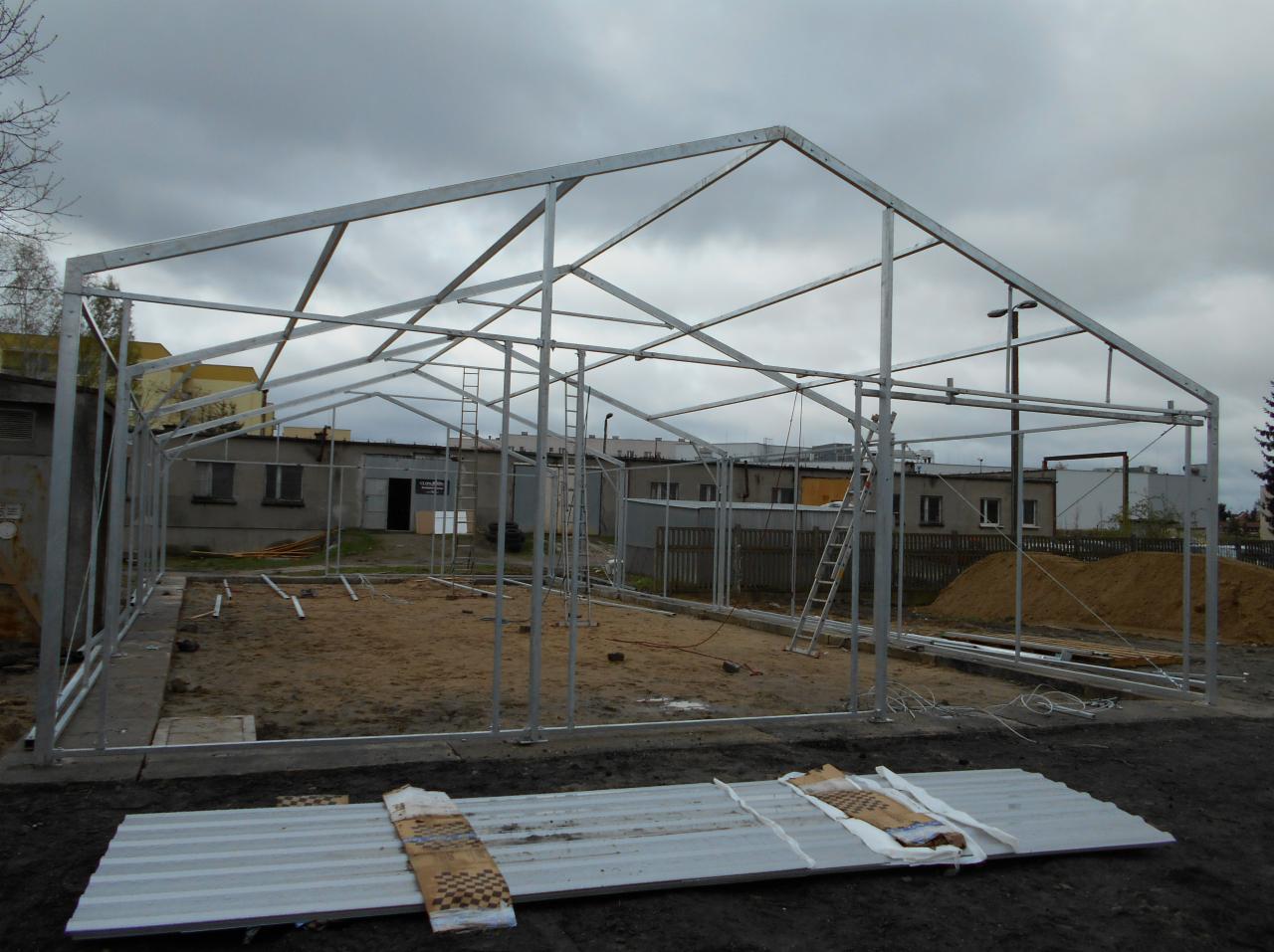 Stalowy szkielet hali w trakcie montażu, widoczne płyty warstwowe w dolnej części kadru, drabina oparta o konstrukcję, pochmurne niebo w tle.