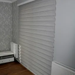 Szara, pozioma przesłona okienna z naprzemiennych pasów tkaniny transparentnej i zaciemniającej, zamontowana w nowoczesnym wnętrzu z szarą tapetą w ornamenty i drewnianą podłogą.