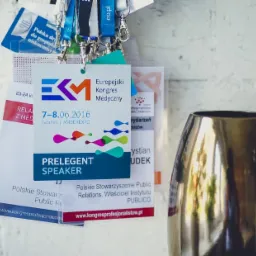 Smycze i identyfikatory uczestników Europejskiego Kongresu Medycznego EKM w Gdańsku, AmberExpo, z datą 7-8.06.2016, wiszące na tle białej ceglanej ściany, obok złoty wazon.