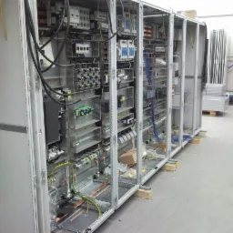 Rozbudowana szafa sterownicza w trakcie montażu, widoczne liczne przewody, moduły i elementy zabezpieczeń, zamontowana w szarej metalowej obudowie na drewnianych podkładkach, w tle inne szafy...