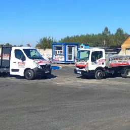 Dwa samochody dostawcze z reklamą firmy oferującej kruszywa, kamienie ogrodowe oraz ziemię, stojące na placu z zapasami materiałów budowlanych i kamieni ozdobnych.