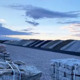 Rzędy usypanych pryzm różnego rodzaju kruszyw, widoczne palety z kostką brukową na pierwszym planie, tło stanowi pochmurne niebo o zmierzchu.