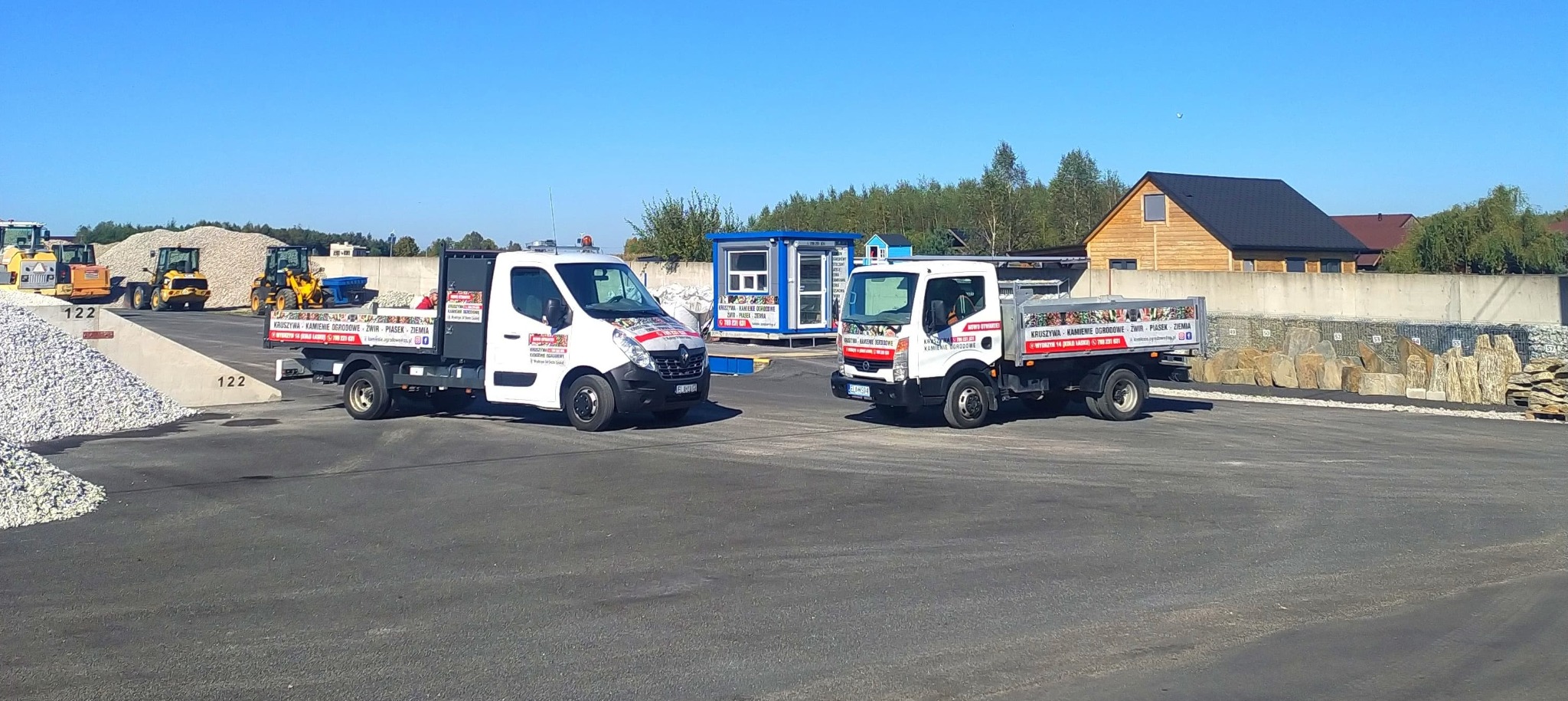 Dwa samochody dostawcze z reklamą firmy oferującej kruszywa, kamienie ogrodowe oraz ziemię, stojące na placu z zapasami materiałów budowlanych i kamieni ozdobnych.