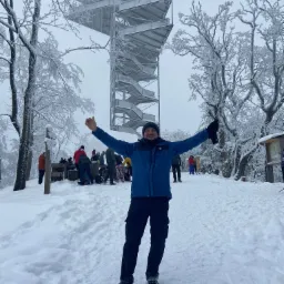 Mężczyzna w zimowej scenerii z rozłożonymi ramionami na tle metalowej wieży widokowej i ośnieżonych drzew