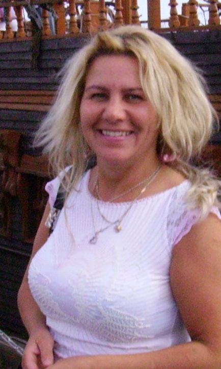 Portret kobiety z blond włosami uśmiechającej się, na tle fragmentu drewnianej konstrukcji przypominającej statek.