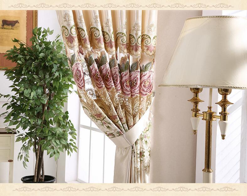 Eleganckie, beżowe zasłony z motywem róż i zielonych ornamentów, upięte kremowym paskiem, widoczne obok lampy stojącej z abażurem i rośliny doniczkowej.