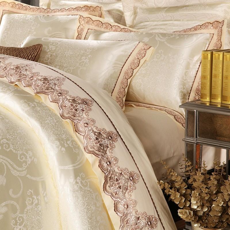 Elegancka pościel w odcieniach kremu i beżu, zdobiona misternym haftem i subtelnym wzorem, częściowo widoczne książki i dekoracja kwiatowa.