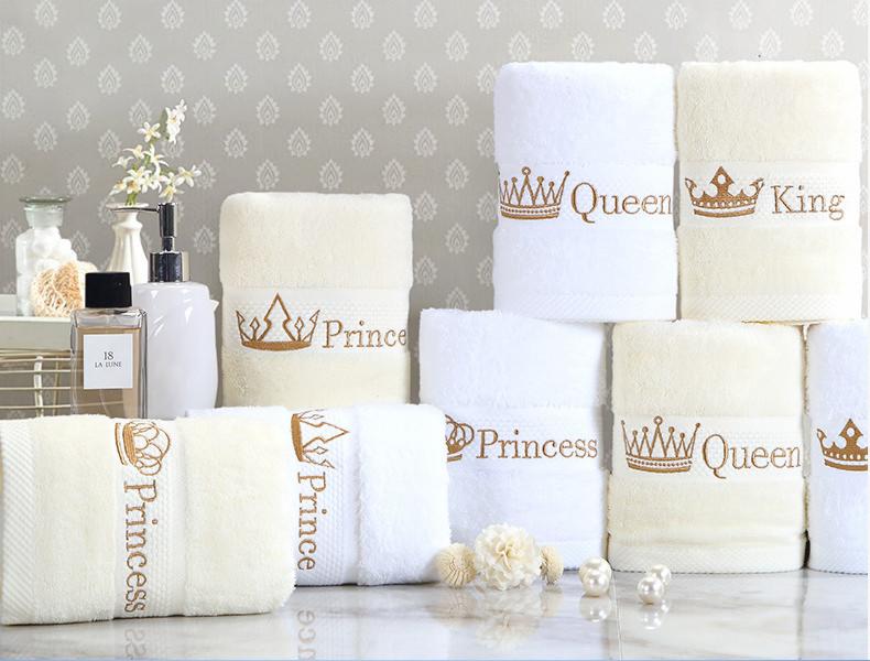 Zestaw ręczników w odcieniach bieli i kremu z haftowanymi koronami i napisami 'King', 'Queen', 'Prince' i 'Princess', ułożone na marmurowym blacie, z flakonem perfum i dozownikiem mydła w tle.