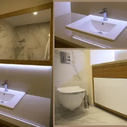 Nowoczesna łazienka z białymi płytkami imitującymi marmur, umywalką nablatową z chromowaną baterią, podświetlanym lustrem w drewnianej ramie oraz szafką pod umywalką z frontami w kolorze drewna...