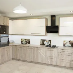 Jasna kuchnia z drewnopodobnymi frontami szafek dolnych i białymi górnymi, z czarnym okapem i dekoracyjnymi panelami szklanymi z motywem kawy.