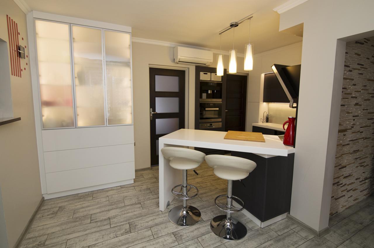 Nowoczesna kuchnia z białym blatem, hokerami na chromowanych nogach i zabudową z matowego szkła, oświetlona wiszącymi lampami, z drewnianą deską do krojenia na blacie.