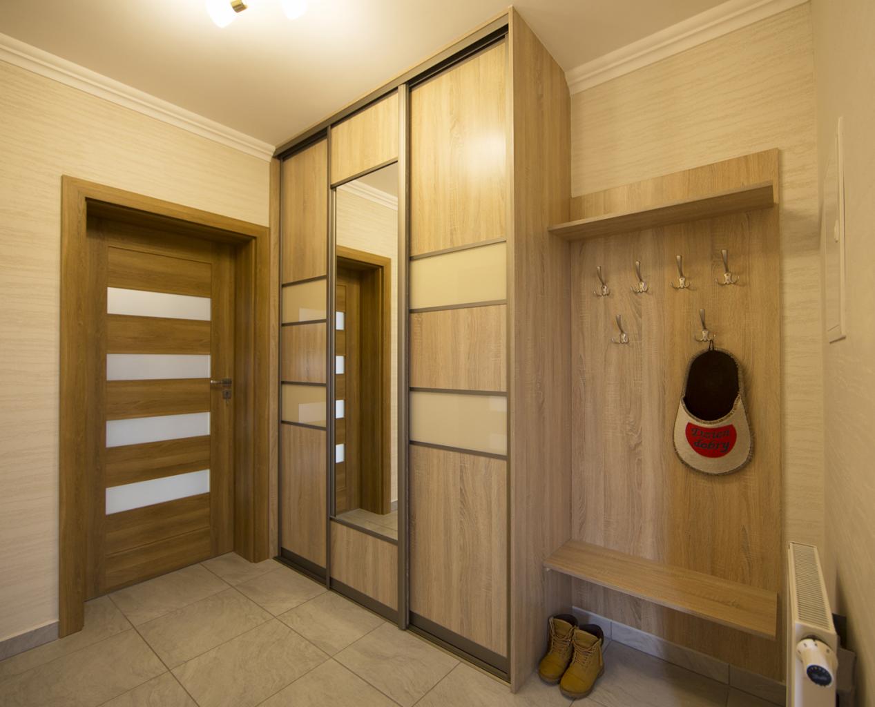Jasny przedpokój z dużą, przesuwną szafą wnękową z lustrem i panelami, wieszakiem z półką, drewnianymi drzwiami z przeszkleniami i parą żółtych butów na podłodze z płytek.