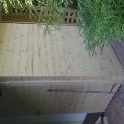 Świeżo wybudowana drewniana szopa z drzwiami, widok z boku, obok leży miotła na kostce brukowej, a nad nią zwisają gałęzie drzewa.