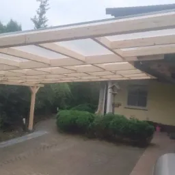 Zadaszenie tarasu z drewnianą konstrukcją i przezroczystym dachem, widok z zewnątrz na tle zieleni.