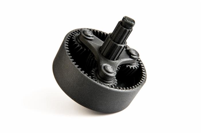 Precyzyjny model przekładni planetarnej, czarny, wykonany metodą addytywną. Widoczne zębatki i oś centralna. Gładka powierzchnia, ostre światło podkreśla detale.