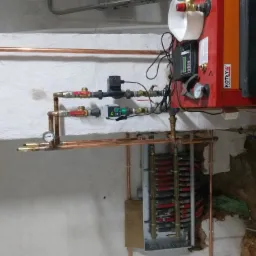 Instalacja grzewcza z piecem Kotly Rakoczy, miedziane rury i zawory, manometry, regulator i rozdzielacz w surowym pomieszczeniu.