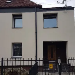 Dwukondygnacyjny budynek z jasną elewacją, ciemnoszarymi akcentami wokół okien i dachu, widoczny zza czarnego, metalowego ogrodzenia; na elewacji widoczny pionowy przewód spustowy.