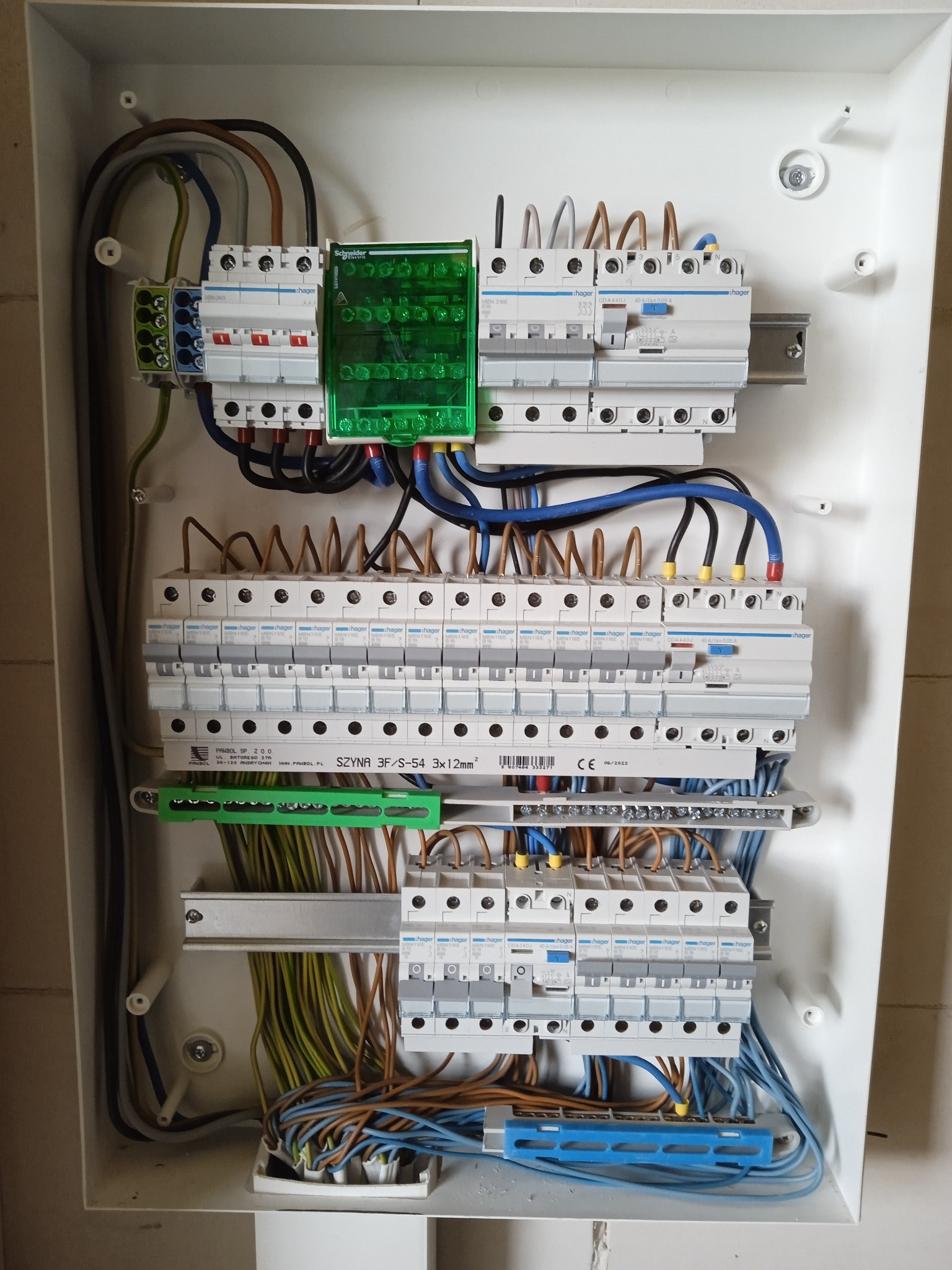 Otwarta skrzynka rozdzielcza z zainstalowanymi bezpiecznikami i przewodami elektrycznymi, widoczne elementy marki Hager i Schneider Electric, szyna montażowa PANBOL.