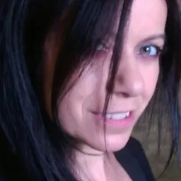 Portret kobiety z długimi, ciemnymi włosami, z jednym pasmem opadającym na twarz, ukazujący jej jasne oczy i lekki uśmiech.