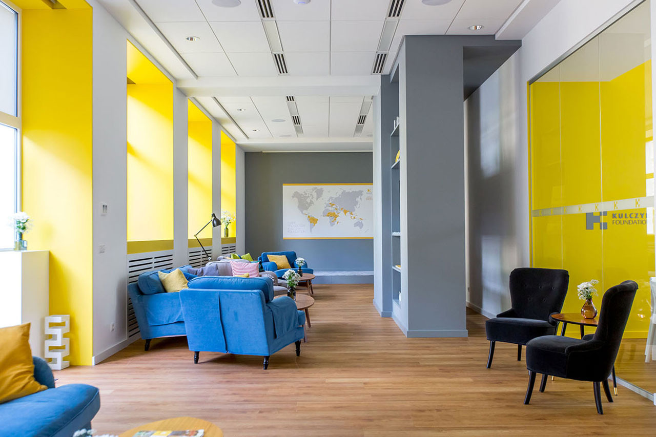 Jasne, przestronne wnętrze biura z żółtymi akcentami, drewnianą podłogą, szarymi ścianami, niebieskimi sofami i czarnymi fotelami, oświetlone światłem dziennym, z widoczną mapą świata na ścianie...