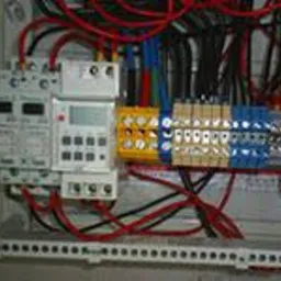 Rozdzielnica elektryczna z widocznymi zabezpieczeniami nadprądowymi, programatorem czasowym i listwami zaciskowymi w kolorach żółtym i niebieskim. Przewody ułożone w wiązki w górnej części obudowy.