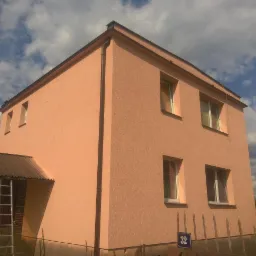 Dwupiętrowy dom z elewacją w odcieniach brzoskwini, widoczny z dołu pod kątem; na elewacji widoczne okna z białymi ramami, brązowe rynny, numer domu 32 na niebieskiej tabliczce, oraz fragment...