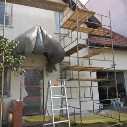 RO-STA Firma Ogólnobudowlana Starosta Roman - Dom w trakcie malowania elewacji: rusztowania, okna i drzwi zabezpieczone folią, podbitka częściowo pomalowana, widoczny fragment dachu z brązową dachówką.