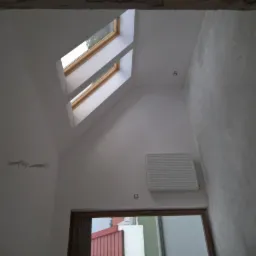 Wnętrze poddasza z dwoma oknami dachowymi, grzejnikiem i ścianą w trakcie wykańczania, widok na zewnątrz przez okno w dole.