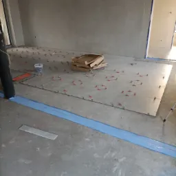 Układanie jasnych płytek podłogowych z użyciem systemu poziomowania, widoczne krzyżyki dystansowe, wiaderko z klipsami i kartonowe opakowanie płytek w surowym wnętrzu z szarymi ścianami...