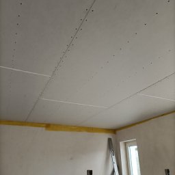 RO-STA Firma Ogólnobudowlana Starosta Roman - Sufit pokryty płytami kartonowo-gipsowymi, widoczne łączenia i ślady po wkrętach, żółta taśma zabezpieczająca przy ścianie, fragment rusztowania i drabina oparta o ścianę z oknem.