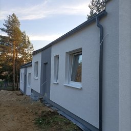 RO-STA Firma Ogólnobudowlana Starosta Roman - Nowo otynkowany dom jednorodzinny z szarą stolarką okienną i grafitowymi rynnami, widok z boku na nierówny teren.