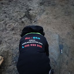 RO-STA Firma Ogólnobudowlana Starosta Roman - Pracownik firmy ogólnobudowlanej w kurtce z logo i numerem telefonu, w trakcie pracy na placu budowy, widok z tyłu.