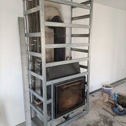 RO-STA Firma Ogólnobudowlana Starosta Roman - Konstrukcja z profili stalowych pod zabudowę kominka z podłączonym przewodem kominowym, widoczne rury instalacyjne i poziomica, w tle biała ściana, na podłodze narzędzia i materiały budowlane.